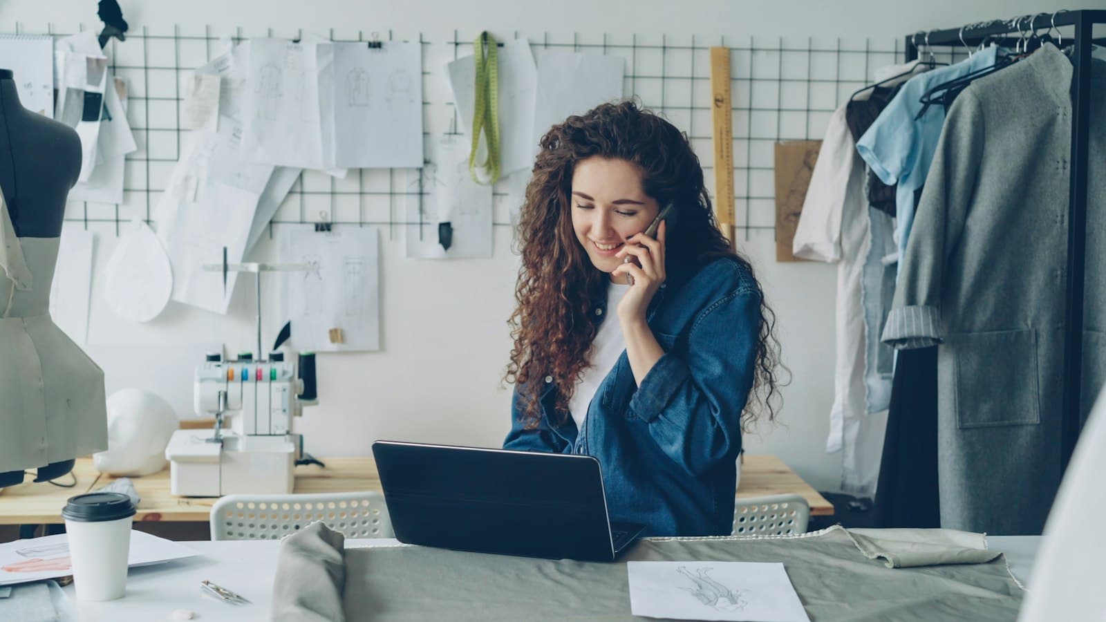 A clothing designer on a phone call at a laptop in a sewing studio, sketches pinned to the wall behind